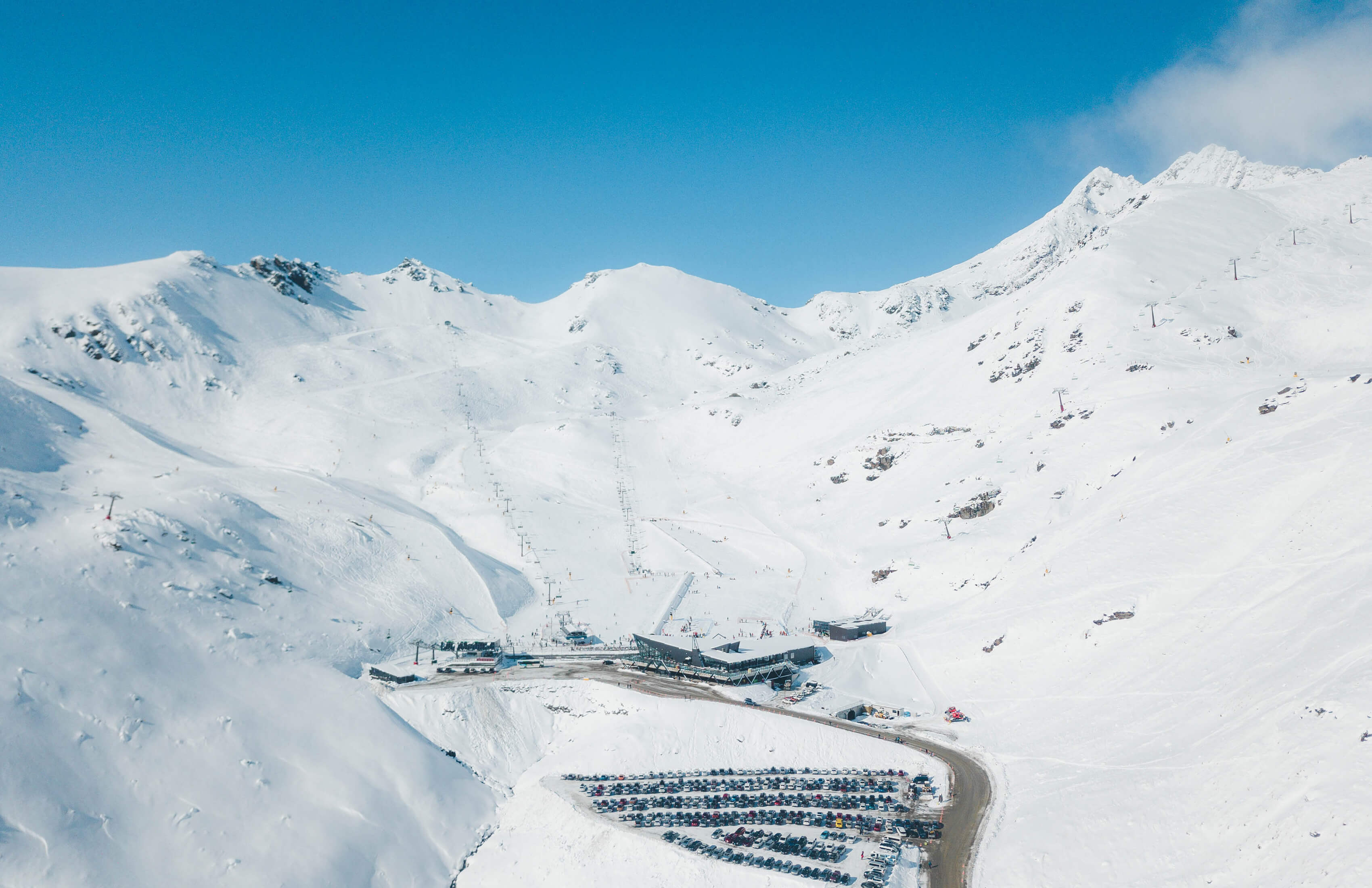 Aerial Shot of a Snow-covered Remarkables ski area with ski lifts, the base building, and a large parking lot filled with cars under a clear blue sky.