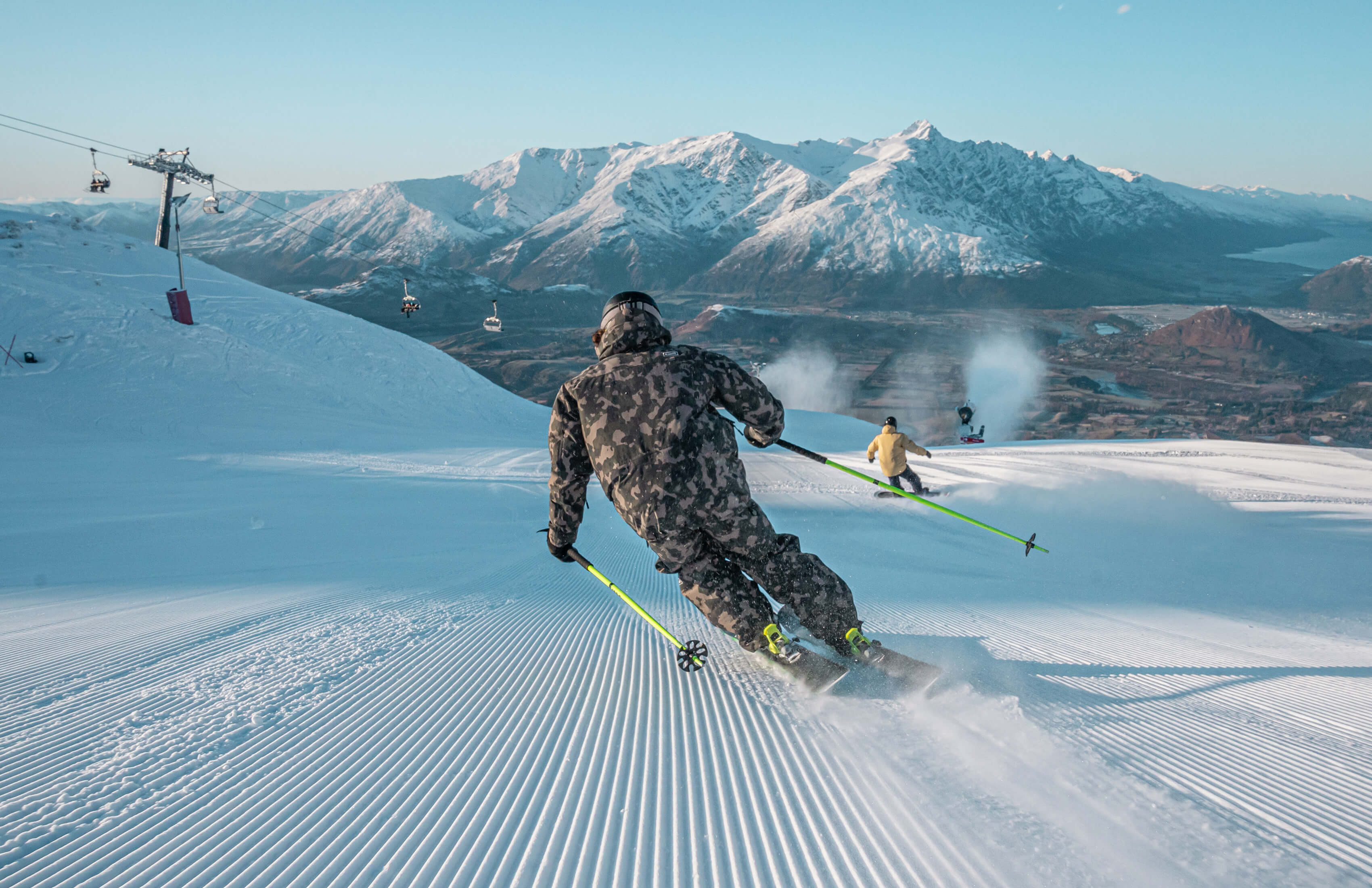 Two skiers descending a freshly groomed ski trail at Coronet Peak with mountains and ski lifts in the background on a clear day.