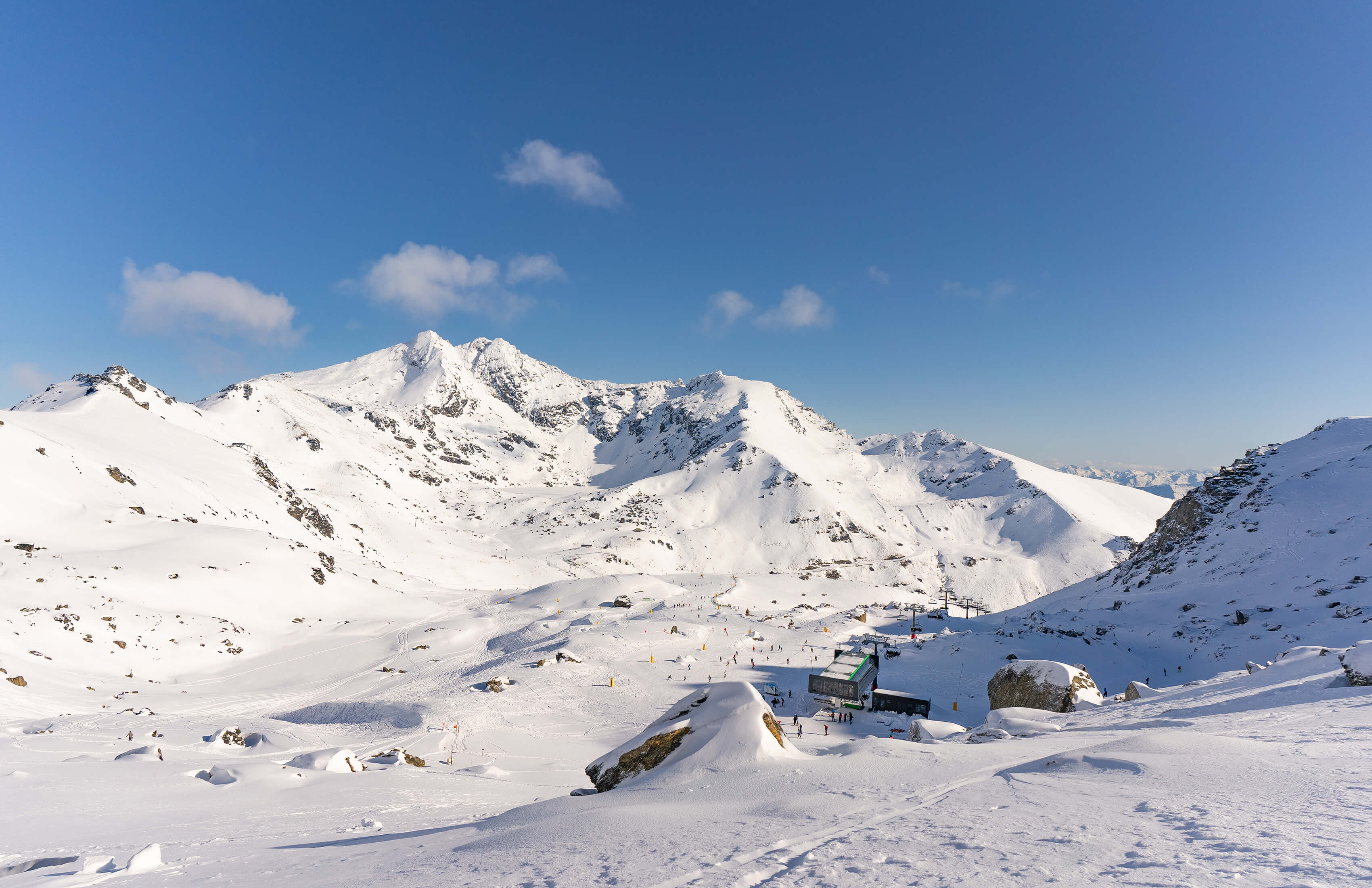 A wide open trail at The Remarkables featuring ski lifts, scattered skiers, and large snow-covered rocks in the foreground.