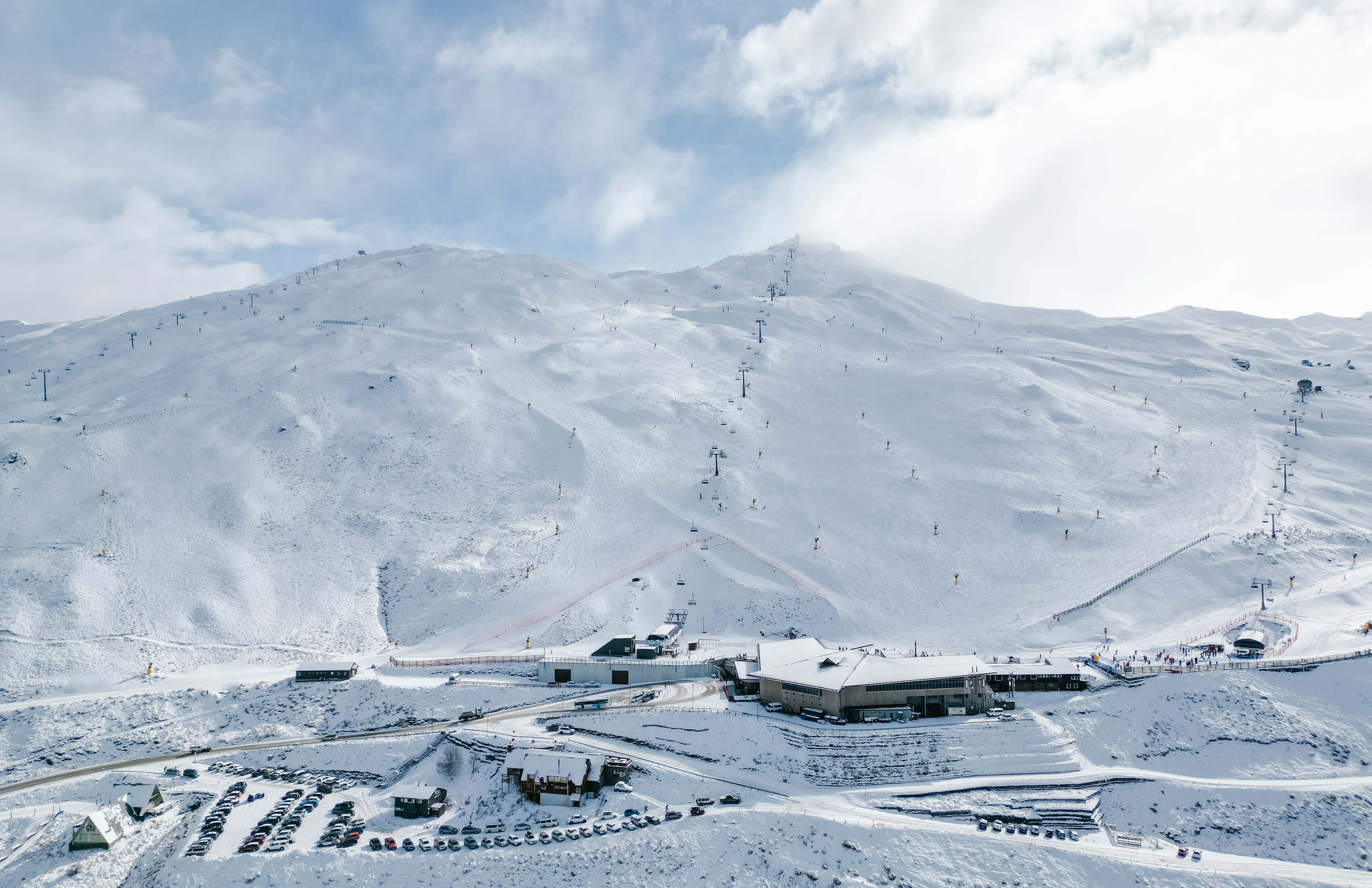 Aerial view of The Remarkables ski resort with ski lifts running up the slopes and a parking area filled with cars in front of the base building.