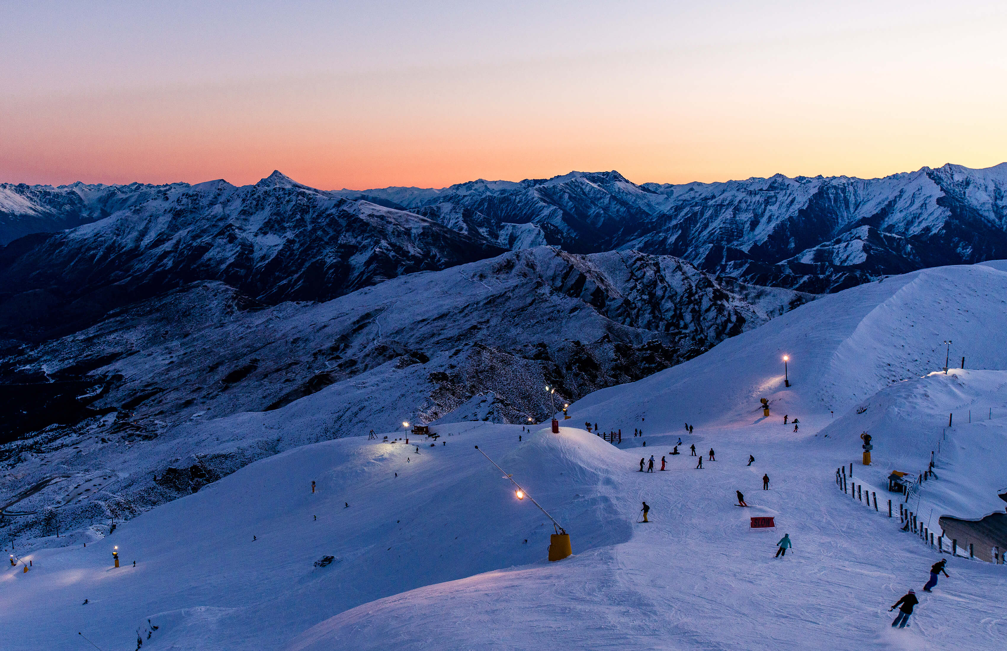 Skiers descend a snow-covered Coronet Peak slope at dusk under lights and a pink-orange sky.