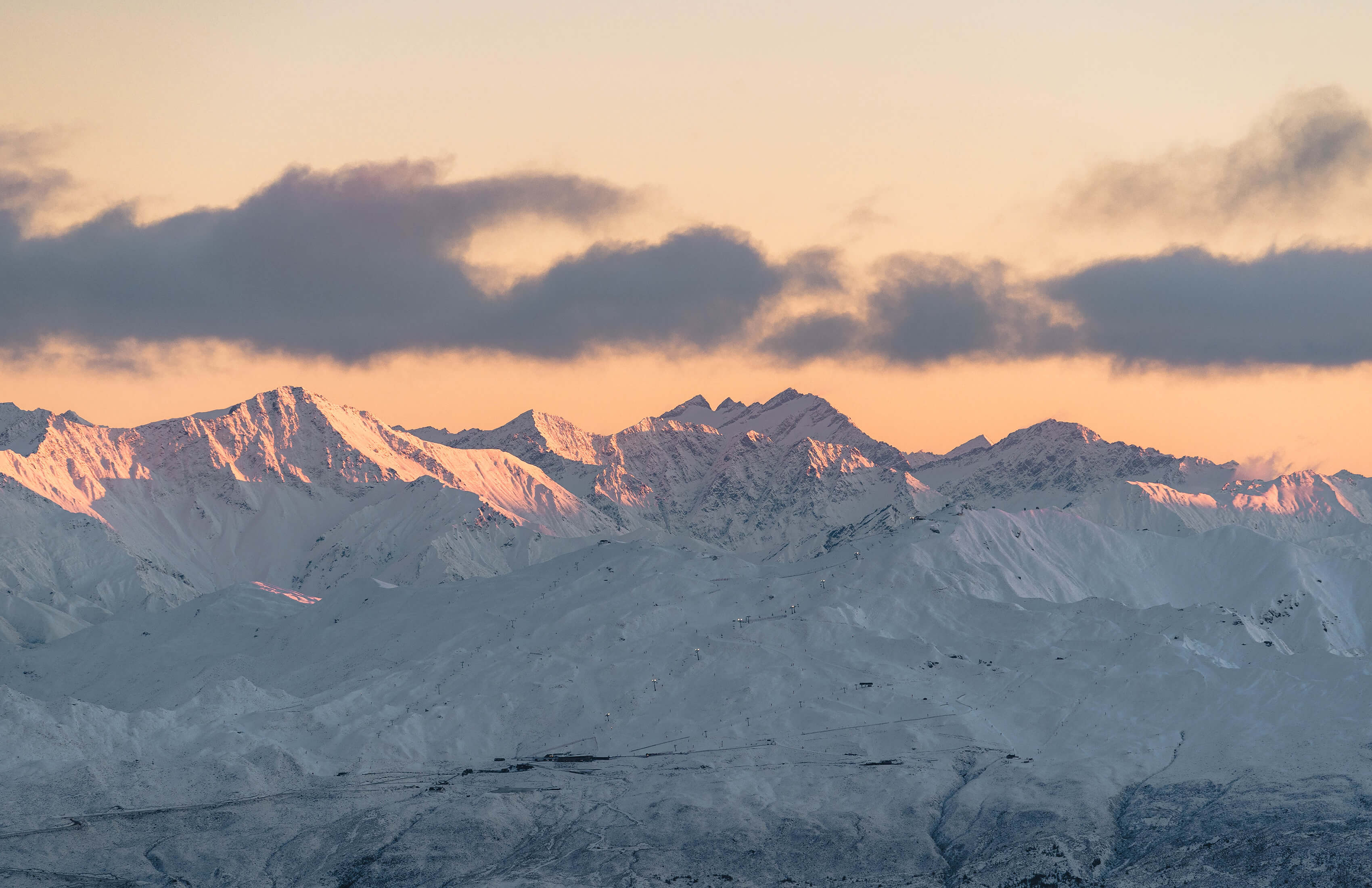Snow-covered Coronet Peak Mountain range at sunrise with soft pink light illuminating the peaks under a partly cloudy sky.